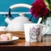 A 380ml Fine China Cat Mug featuring a delightful feline illustration and floral motif sits gracefully on a wooden table next to a white plate filled with macarons and cookies. A teapot and a blurred vase with flowers add an elegant touch to the scene against the blue-tiled wall.