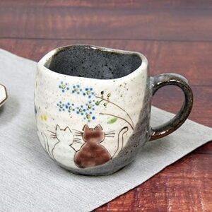 Introducing the "Coffee Mug Sunny Place," a ceramic mug adorned with a hand-painted design of two cats relaxing beneath blue flowers on a textured background. It features an earthy, speckled finish and a rounded handle, making it ideal for savoring your coffee in a bright spot.