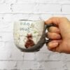 A hand holds the Coffee Mug Sunny Place, adorned with a painted design of two cats sitting together, looking at a tree with blue flowers. The charming scene contrasts beautifully against the white brick wall in the background.