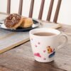 A Colour Changing Mug featuring vibrant cat and paw print designs is filled with tea and rests on a rustic wooden table. Behind it, a plate holds two donuts—one adorned with chocolate frosting—beside a wooden chair.